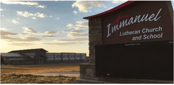 A large sign for Immanuel Lutheran Church and School stands in the foreground, illuminated by LED parking lot lights; in the background, buildings sit under a partly cloudy sky at sunset.