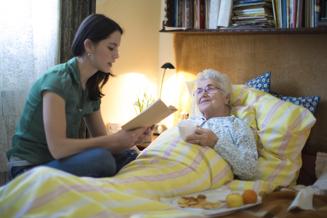 A woman reads to an elderly woman in bed, holding a cup.
