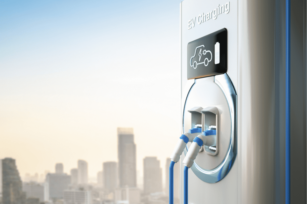 A close-up of an electric vehicle charging station with connectors plugged in. The modern exterior features a screen displaying an icon of a car and the words "EV Charging." In the background, there is a blurred city skyline under a clear sky, hinting at the integration of v2g technology.