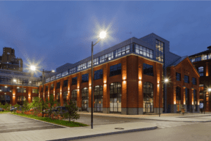 A modern, industrial-style building with illuminated windows at dusk, featuring red brick architecture, large street lamps, and a mix of greenery and paved areas in the foreground. Ev chargers are subtly integrated into the landscape. The sky is dark blue, indicating early evening.