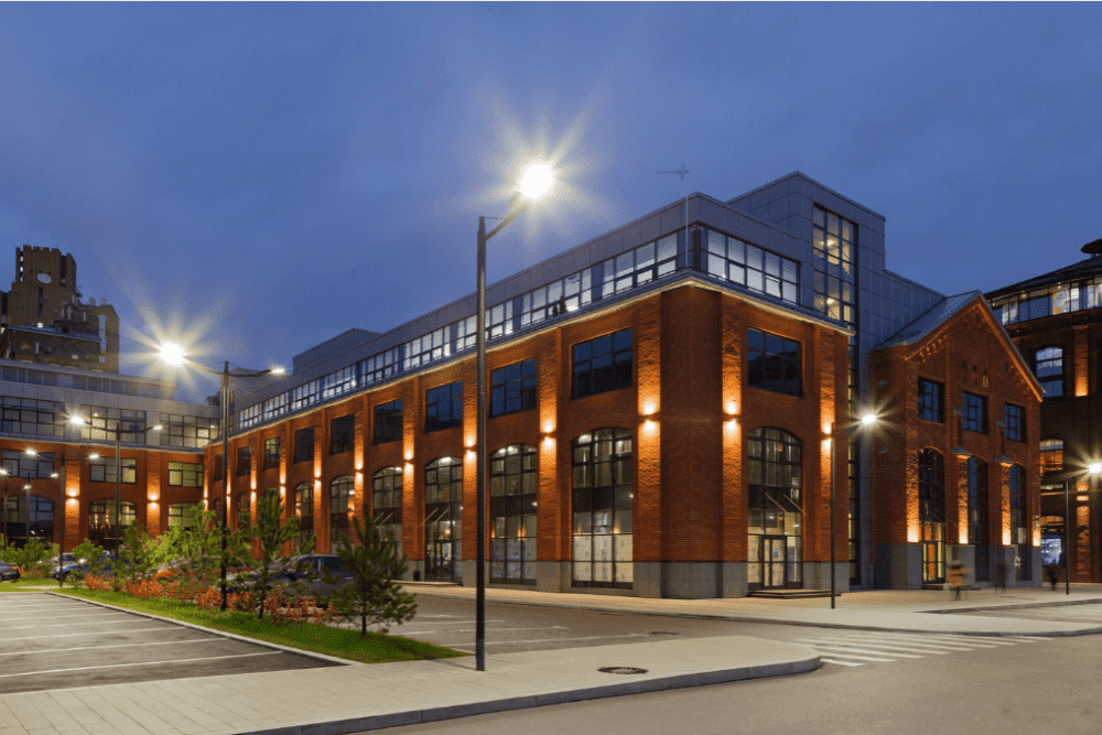 A modern, industrial-style building with illuminated windows at dusk, featuring red brick architecture, large street lamps, and a mix of greenery and paved areas in the foreground. Ev chargers are subtly integrated into the landscape. The sky is dark blue, indicating early evening.