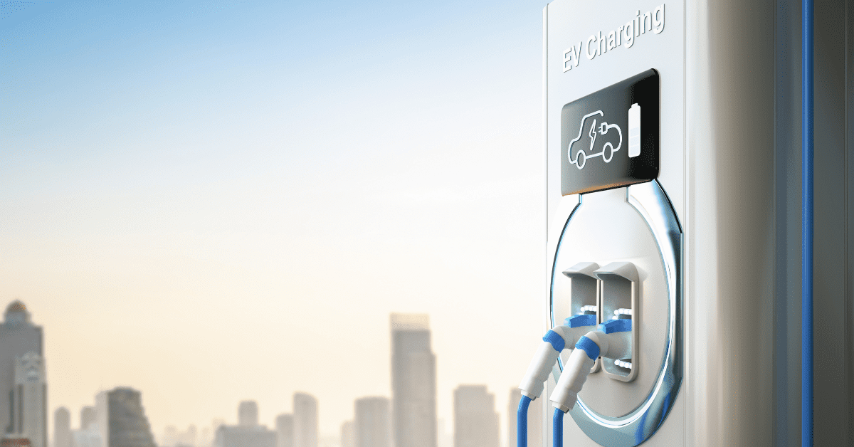 A close-up of an electric vehicle (EV) charging station with two cables plugged in. The background features a blurred urban skyline with tall buildings under a clear blue sky. The station is marked with "EV Charging" and an icon of a charging car, showcasing cutting-edge V2G technology.