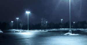 A dimly lit parking lot at night comes alive with strategic parking lot lighting solutions, as several light poles illuminate the area. Patches of snow are scattered along the edges of the asphalt. Bare trees line the background under a dark, cloudy sky.