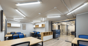 A modern office interior with wooden desks and blue mesh chairs. The room features white walls, overhead fluorescent lighting, and a glass door leading to another office area. Shelves and a clock are mounted on the walls.