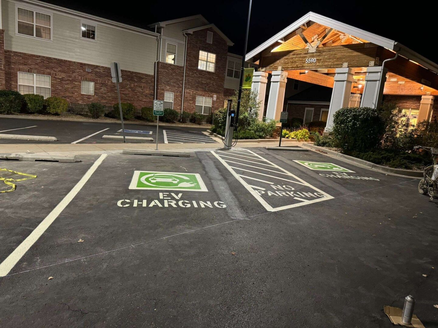 Night view of an EV charging station with two designated parking spots. One spot is marked for "EV Charging" and the other as "No Parking." A building entrance with lit windows is visible in the background.