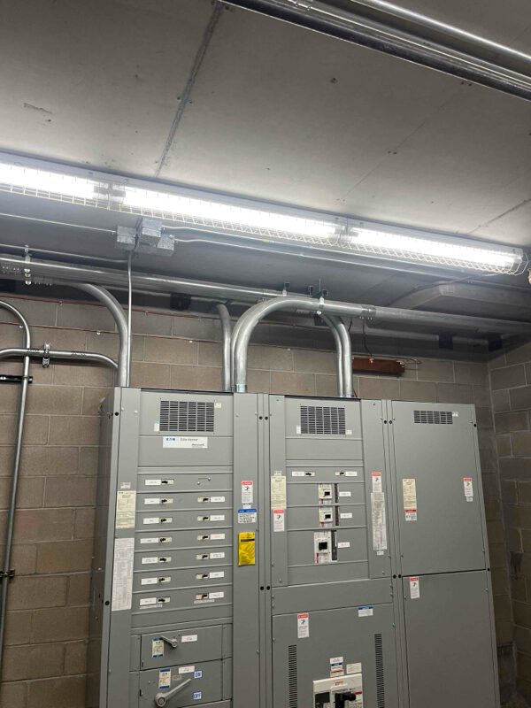 A dimly lit electrical room with gray control panels and circuit breakers mounted on a brick wall. Overhead fluorescent lights illuminate the space, and several metal conduits run from the panels to the ceiling.