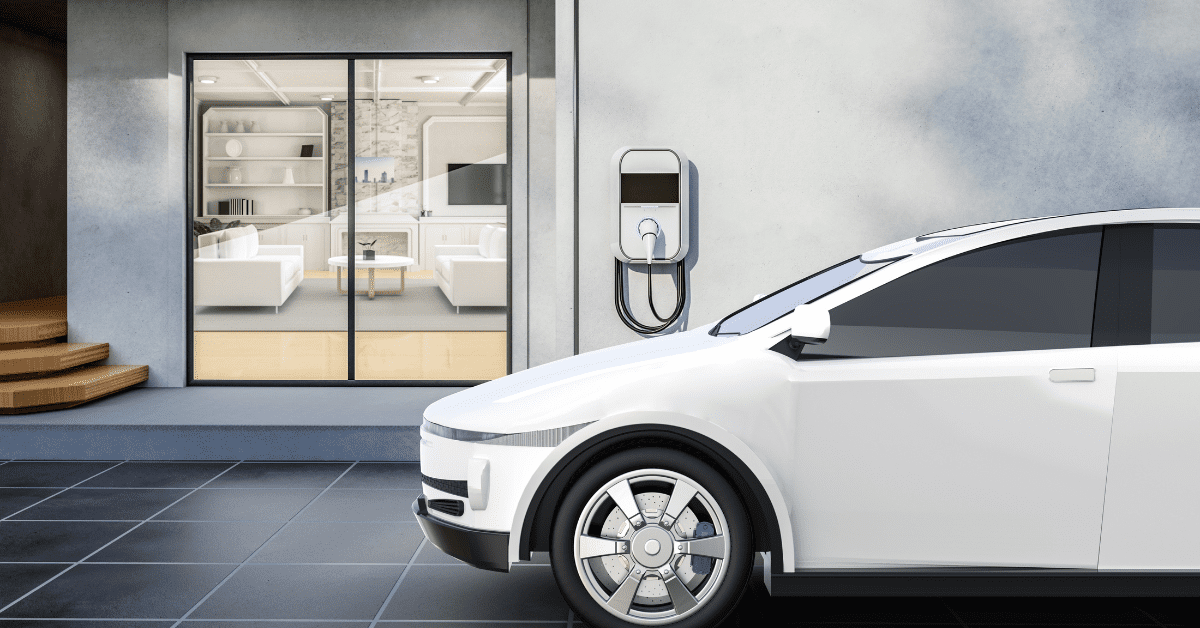 A white electric car is parked outside a modern home, connected to a wall-mounted charging station. Large glass doors reveal a stylish interior with white furniture and wooden accents.