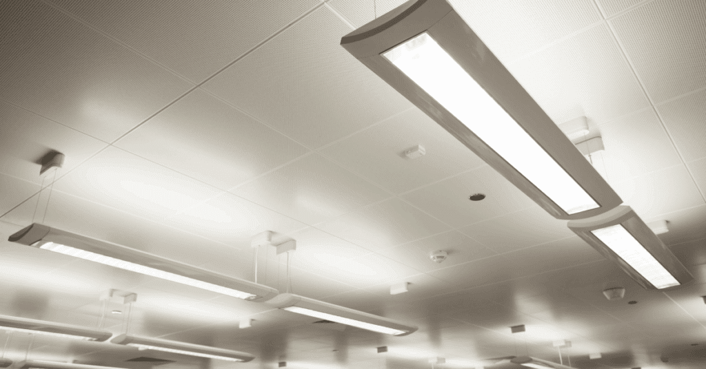 Close-up view of a white, modern office ceiling with several rectangular fluorescent light fixtures, ceiling tiles, air vents, and smoke detectors. The lights are brightly illuminated.