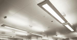 Close-up view of a white, modern office ceiling with several rectangular fluorescent light fixtures, ceiling tiles, air vents, and smoke detectors. The lights are brightly illuminated.