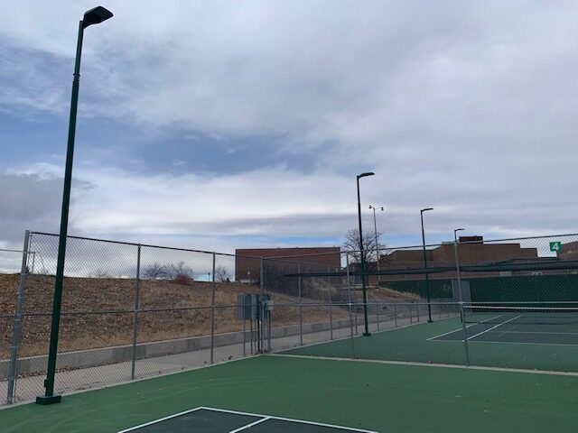 An empty outdoor tennis court surrounded by a chain-link fence, with several tall light poles and buildings visible in the background under a cloudy sky.