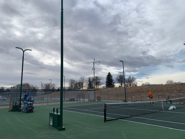 A mostly empty outdoor tennis court under a cloudy sky, with some equipment and lights visible. A person in an orange jacket walks near the fence in the background. Trees and a parking lot are also in the distance.