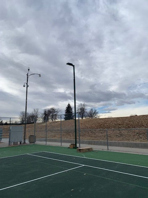 A cloudy sky above an empty outdoor tennis court, surrounded by a chain-link fence and trees. A single light pole stands near the court, with a dirt embankment and sparse vegetation in the background.