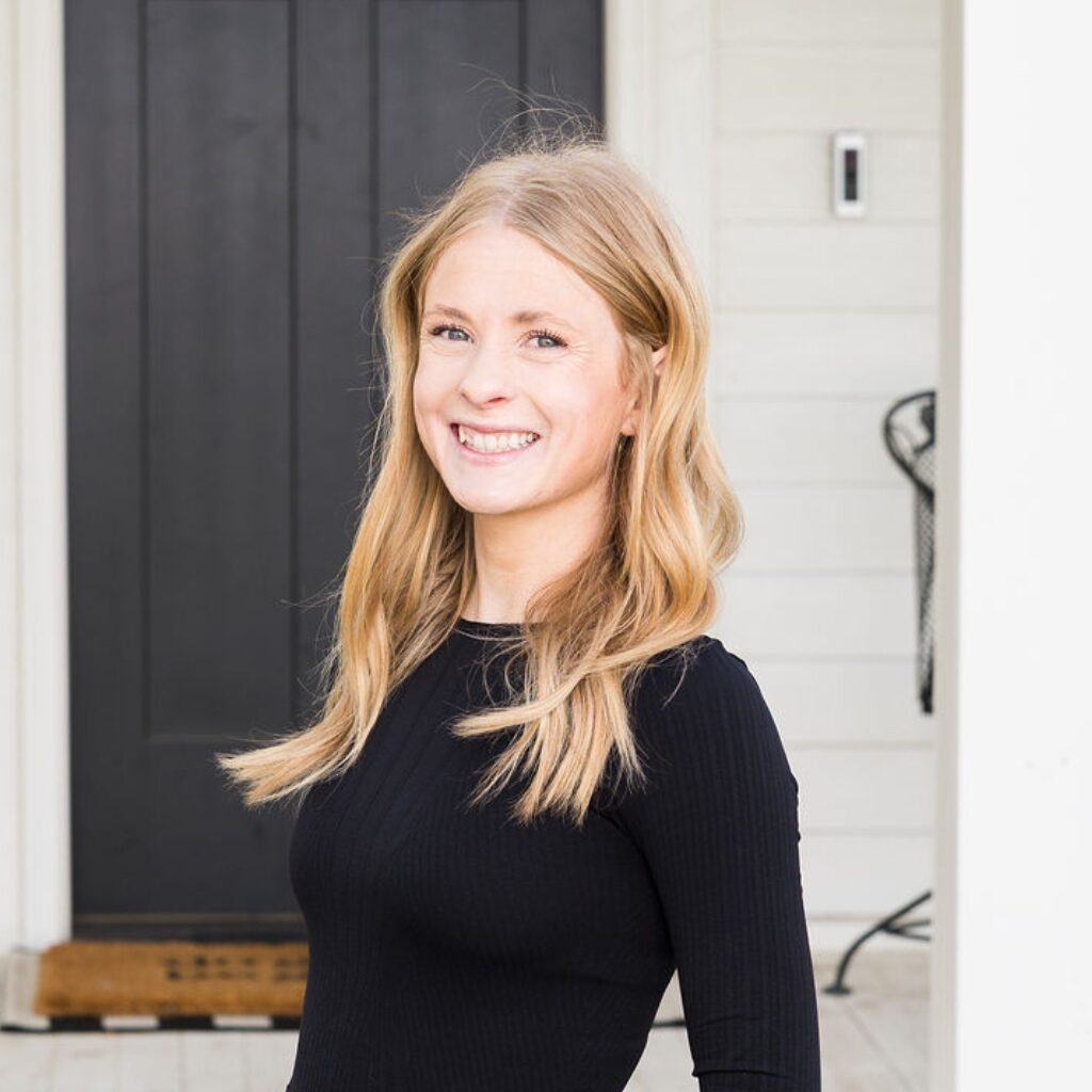 A smiling woman with long blonde hair, wearing a black long-sleeve top, stands in front of a black door and light-colored exterior wall.