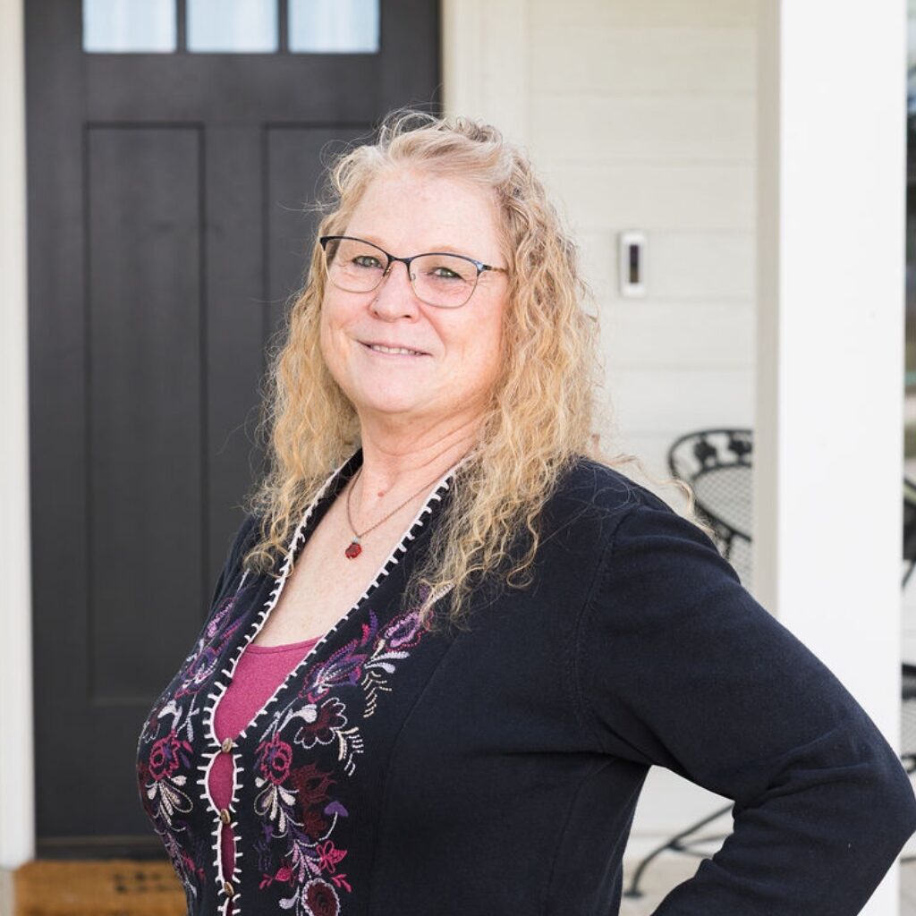 A woman with wavy blonde hair and glasses stands smiling in front of a black door. She wears a dark floral-embroidered cardigan over a maroon top. There is a metal chair and table in the background.