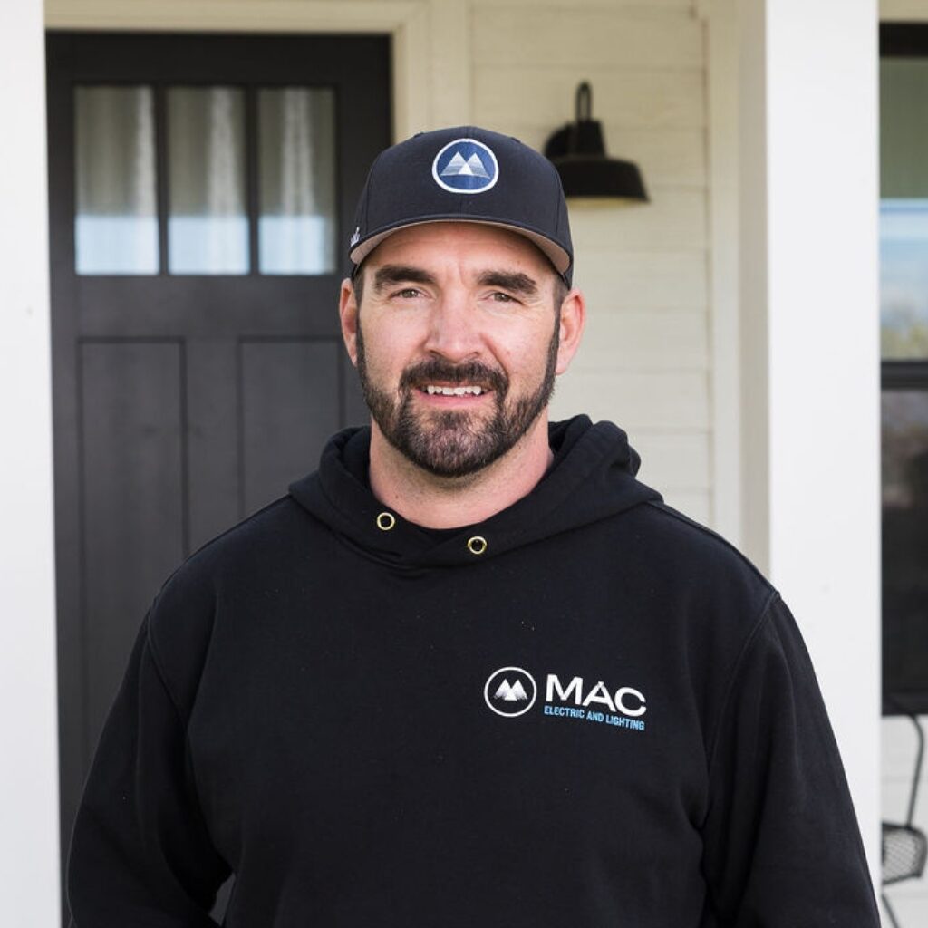 A man with a dark beard, wearing a black cap and a black "MAC Electric and Lighting" hoodie, stands smiling in front of a white house with a black door and window behind him.