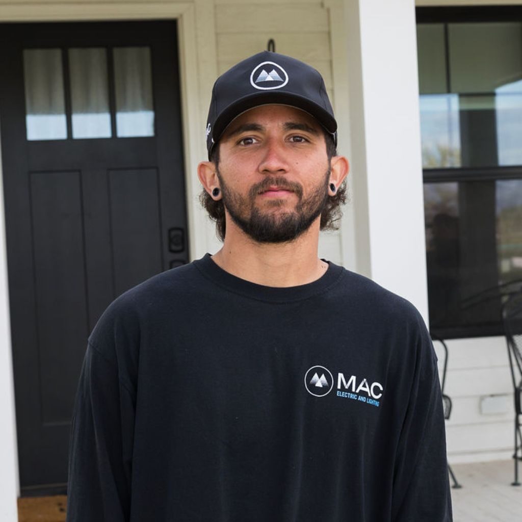 A man with a beard and mustache, wearing a black cap and a black MAC Electric and Lighting shirt, stands in front of a house with a black door and white walls.