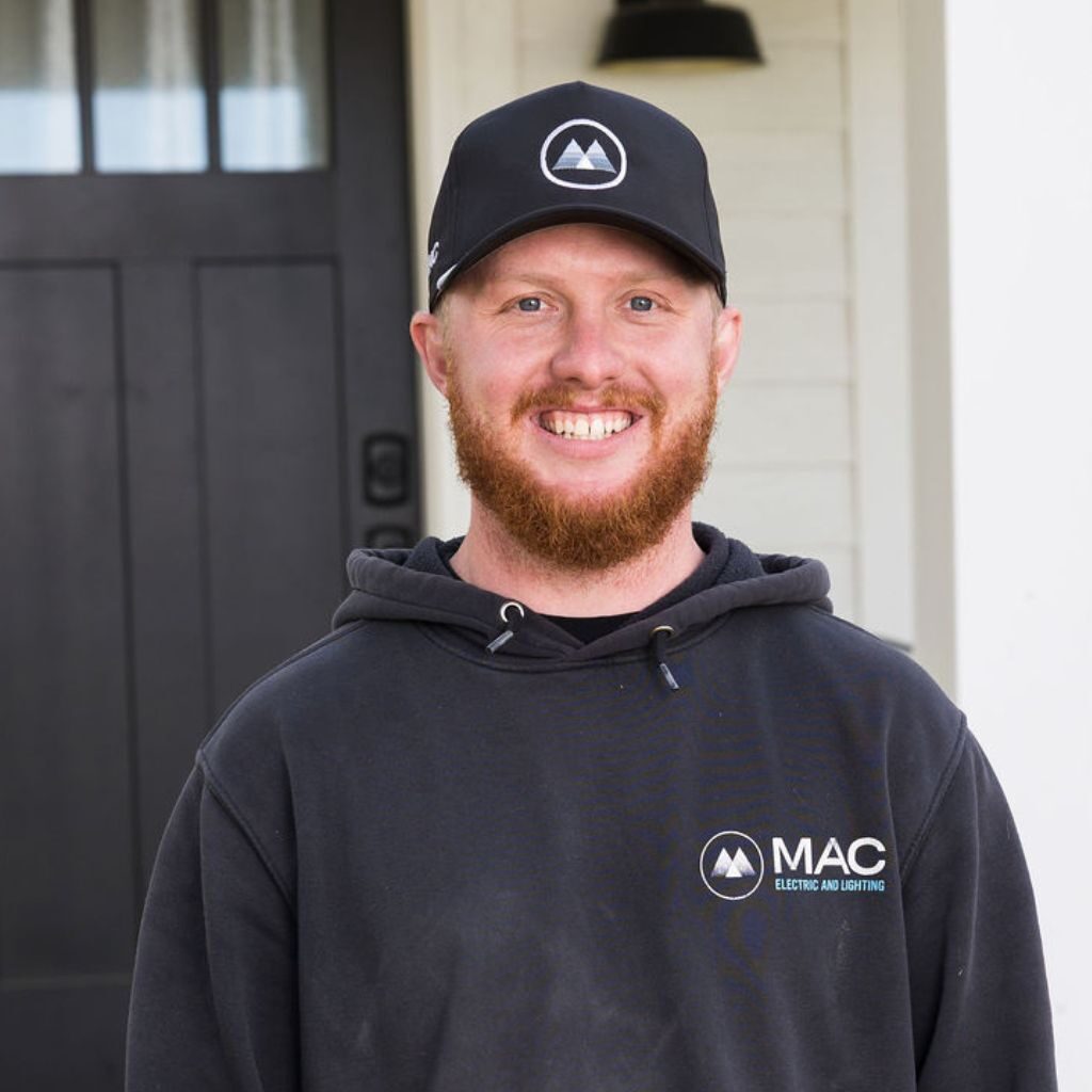 A smiling person with a red beard wears a black cap and hoodie with the "MAC Electric and Lighting" logo, standing in front of a dark door and white wall.