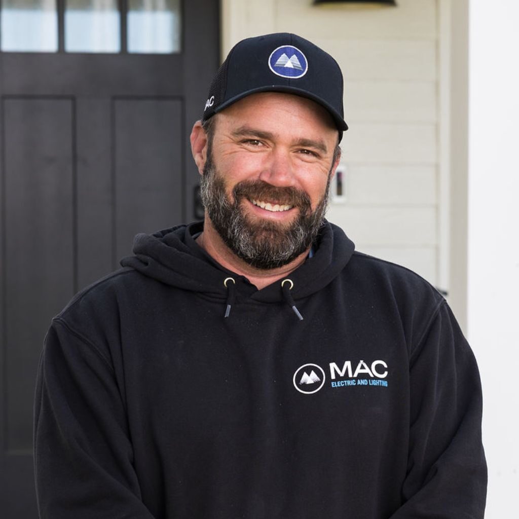 A bearded man in a black cap and black hoodie with "MAC Electric and Lighting" logo stands smiling in front of a black door and white wall.