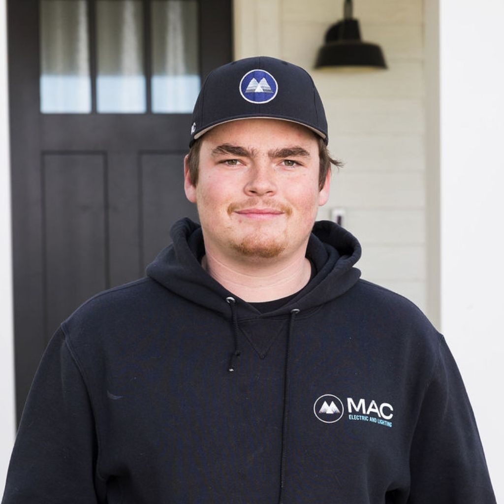 A young man wearing a black cap and a black hoodie with "MAC Electric and Marine" logo stands in front of a dark door and cream-colored wall, looking at the camera with a slight smile.