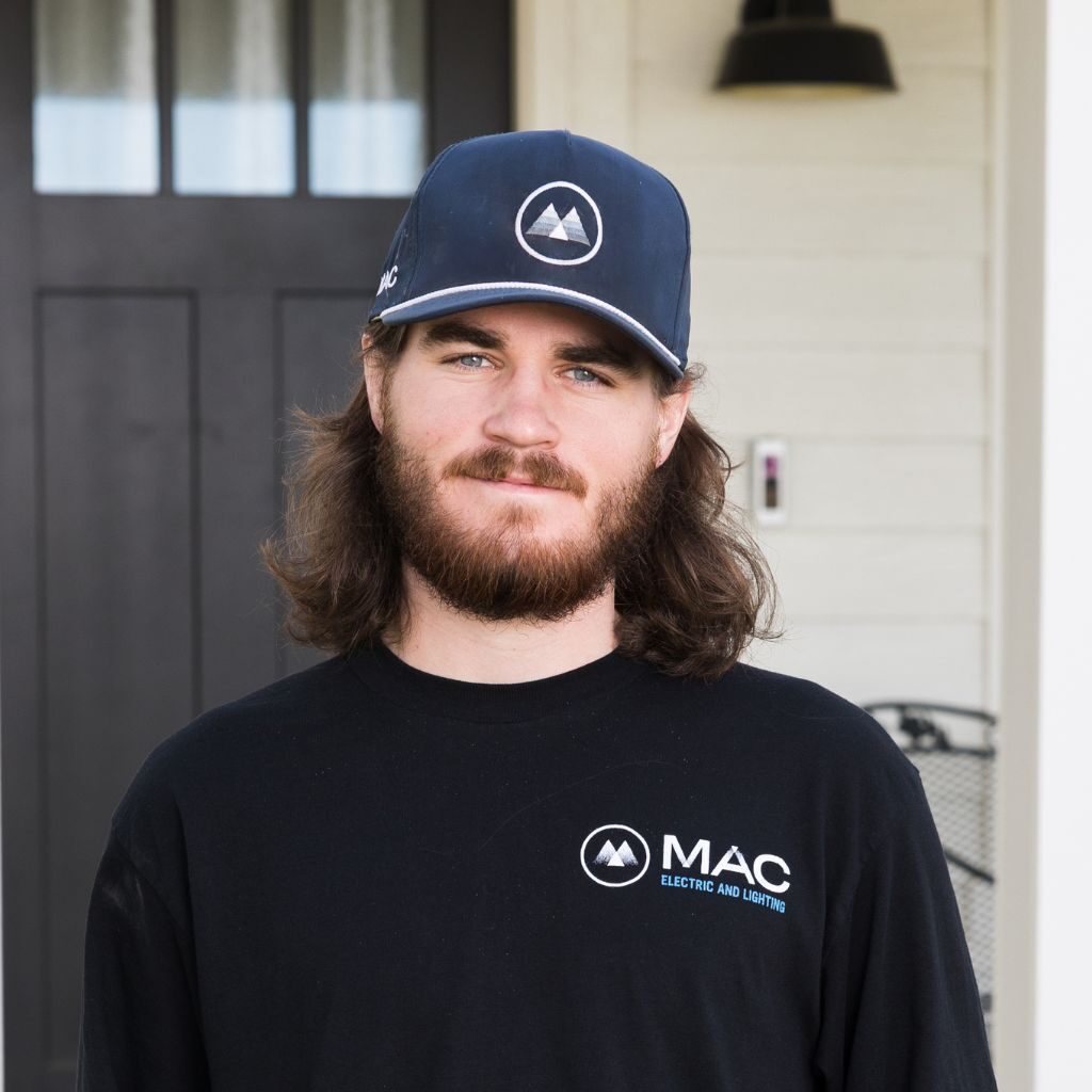 A man with long brown hair and a beard, wearing a navy hat and a black shirt with the "MAC Electric and Lighting" logo, stands in front of a house with a dark door and white siding.