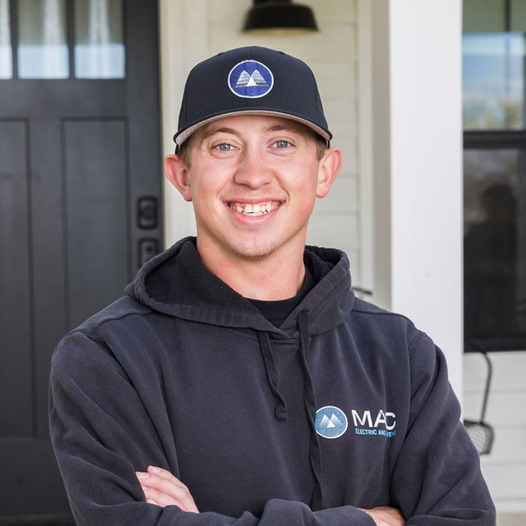 A young man in a black hoodie and cap with the MAC logo stands smiling with arms crossed in front of a black door and white house.