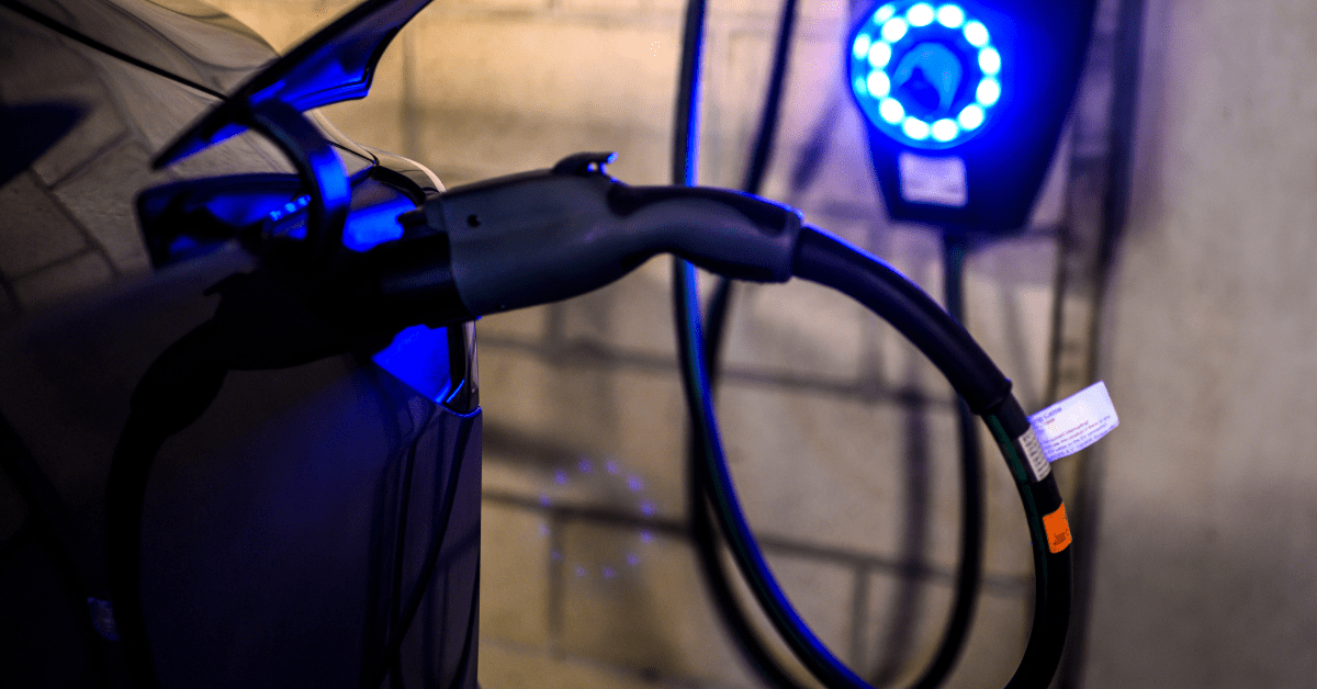 Close-up of an electric vehicle being charged, with a cable plugged in and a blue-lit charging station on the wall—raising the question: Where do EV charging stations get their electricity from?.