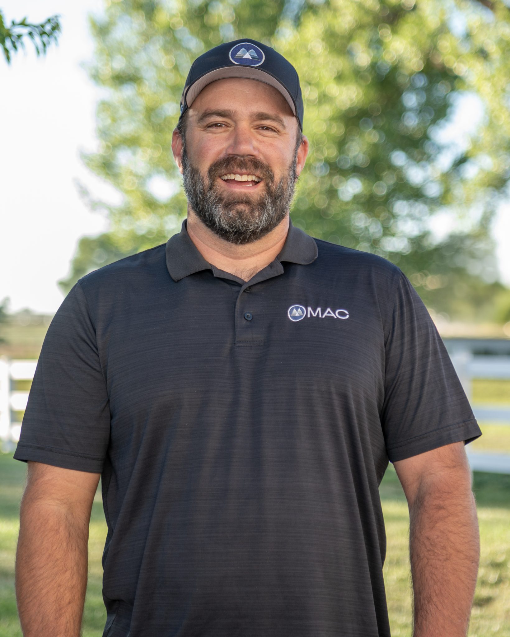 A man wearing a MAC logo polo shirt and cap stands outdoors smiling.