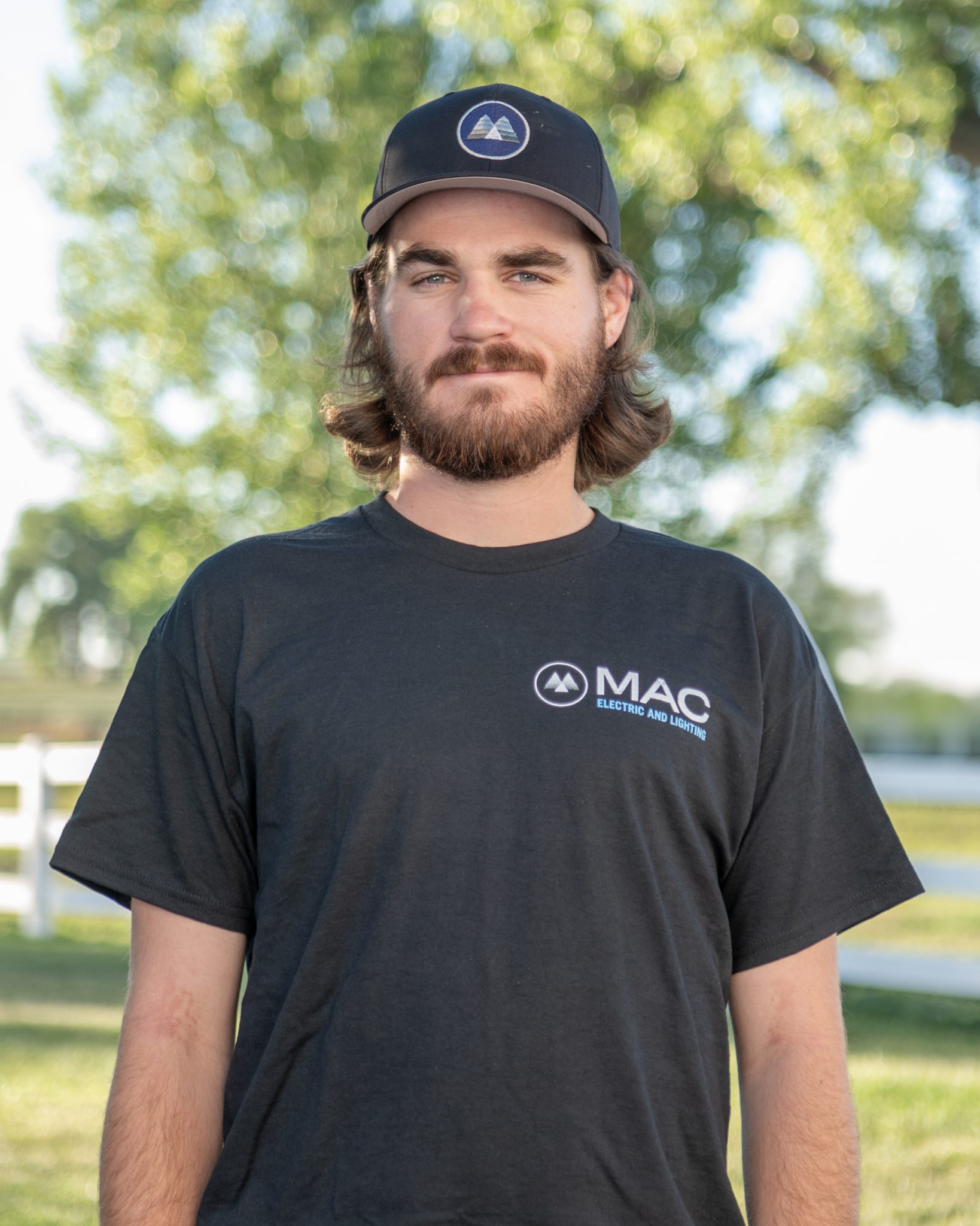 A man wearing a MAC Electric and Lighting t-shirt and a cap stands outdoors.