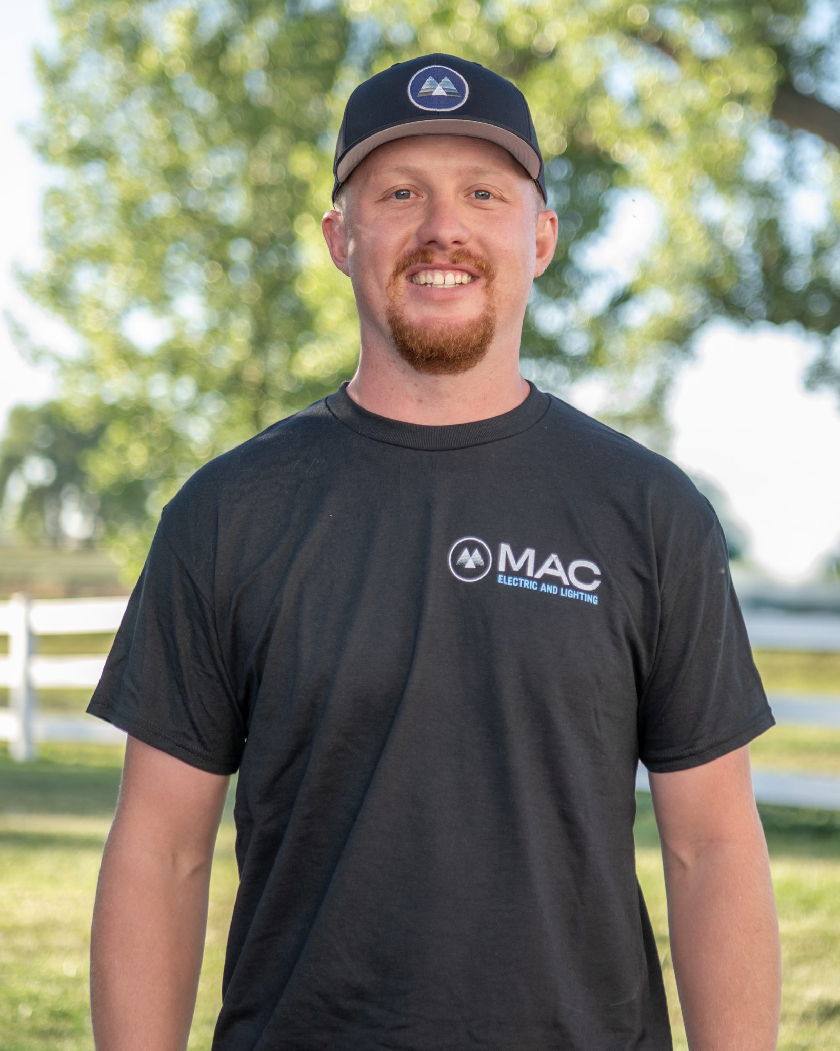 Man in MAC Electric and Lighting shirt outdoors with blurred greenery in the background.