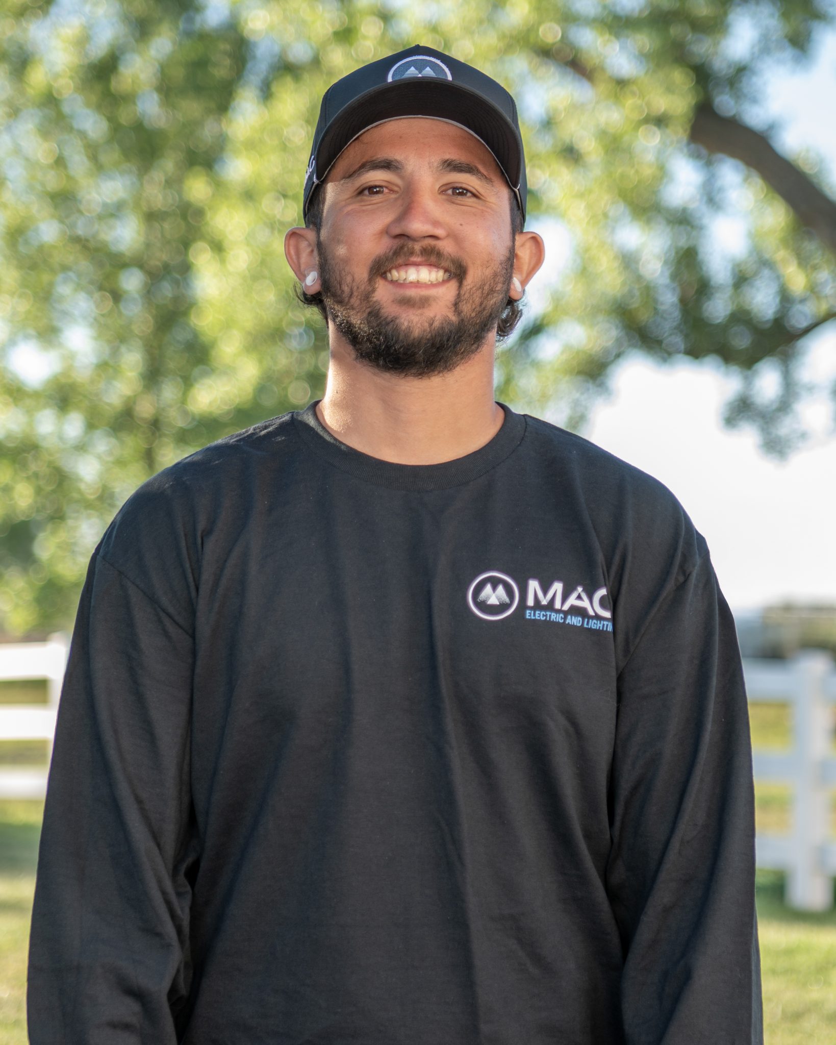 A person smiling outdoors in a black sweatshirt and cap, with trees in the background.