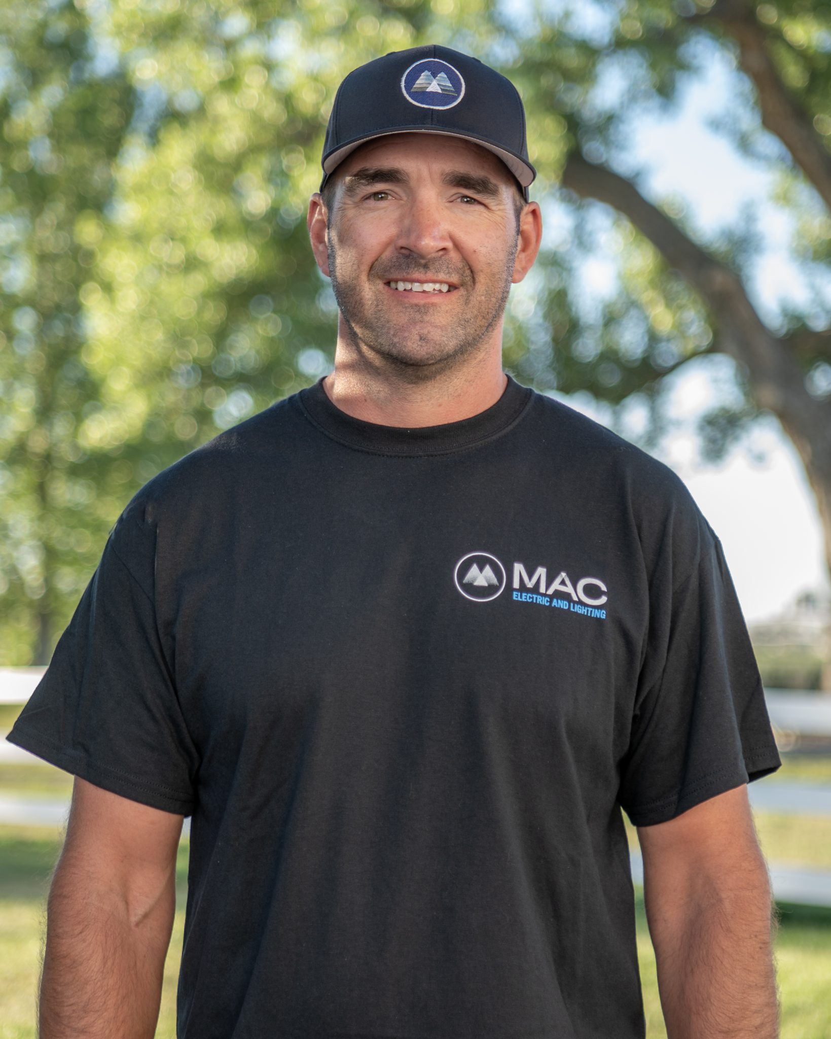 Man in MAC Electric and Lighting t-shirt and cap, smiling outdoors.