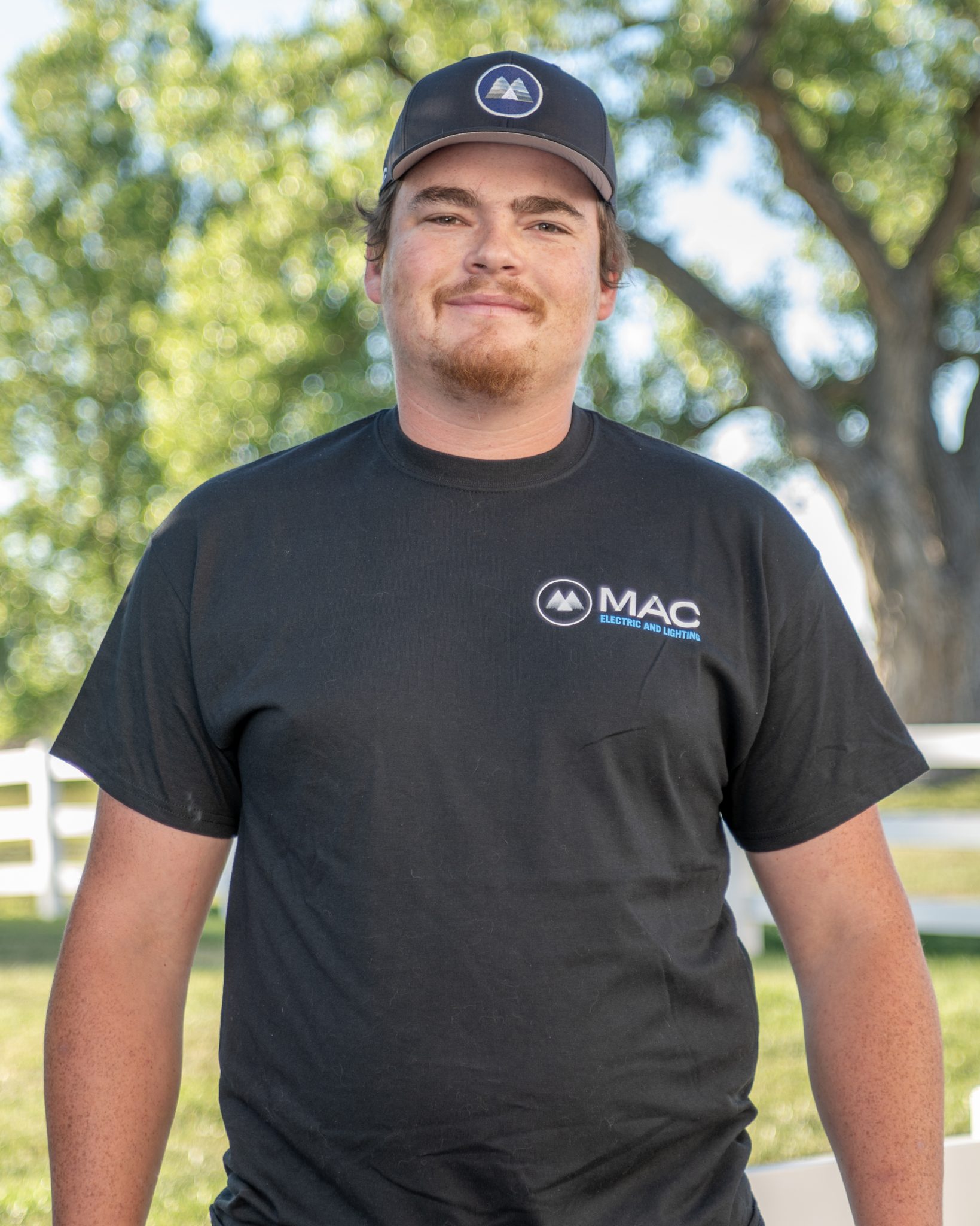 A person in a MAC Electric and Lighting shirt and cap, standing outdoors with trees in the background.
