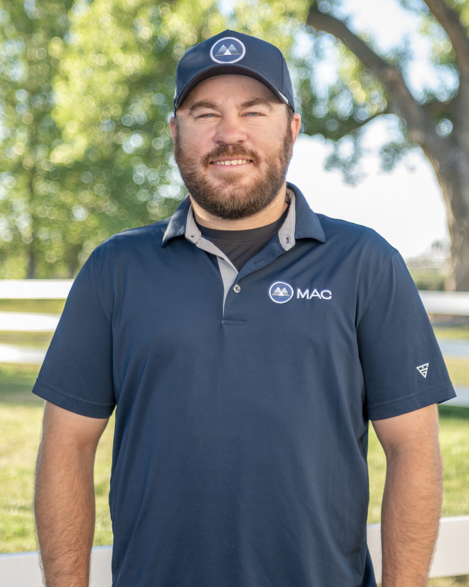 A man in a navy MAC shirt and cap stands outdoors with trees in the background.