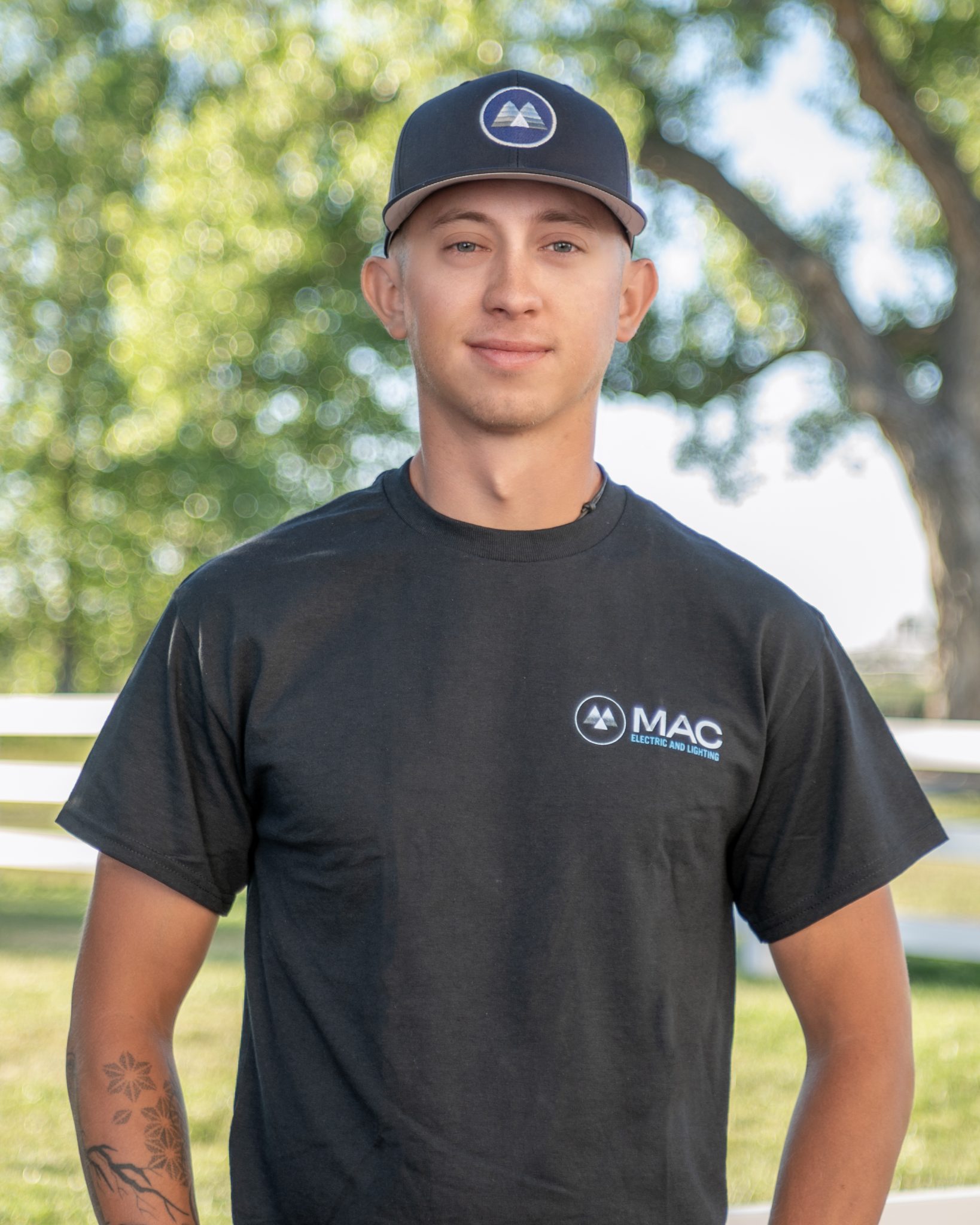 A man in a black MAC Electric and Lighting shirt outdoors with trees in the background.