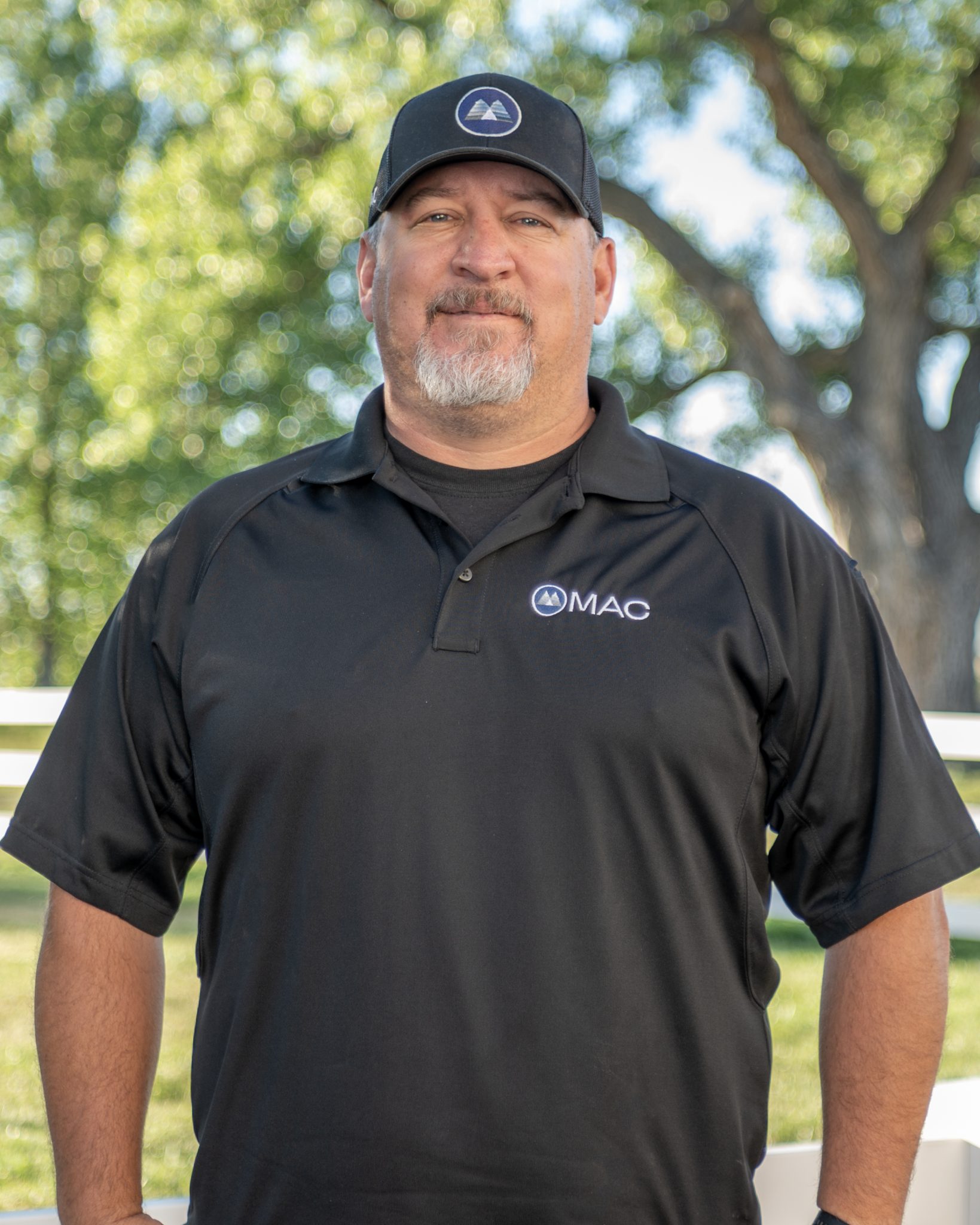 A man in a black MAC polo and cap standing outdoors with trees in the background.