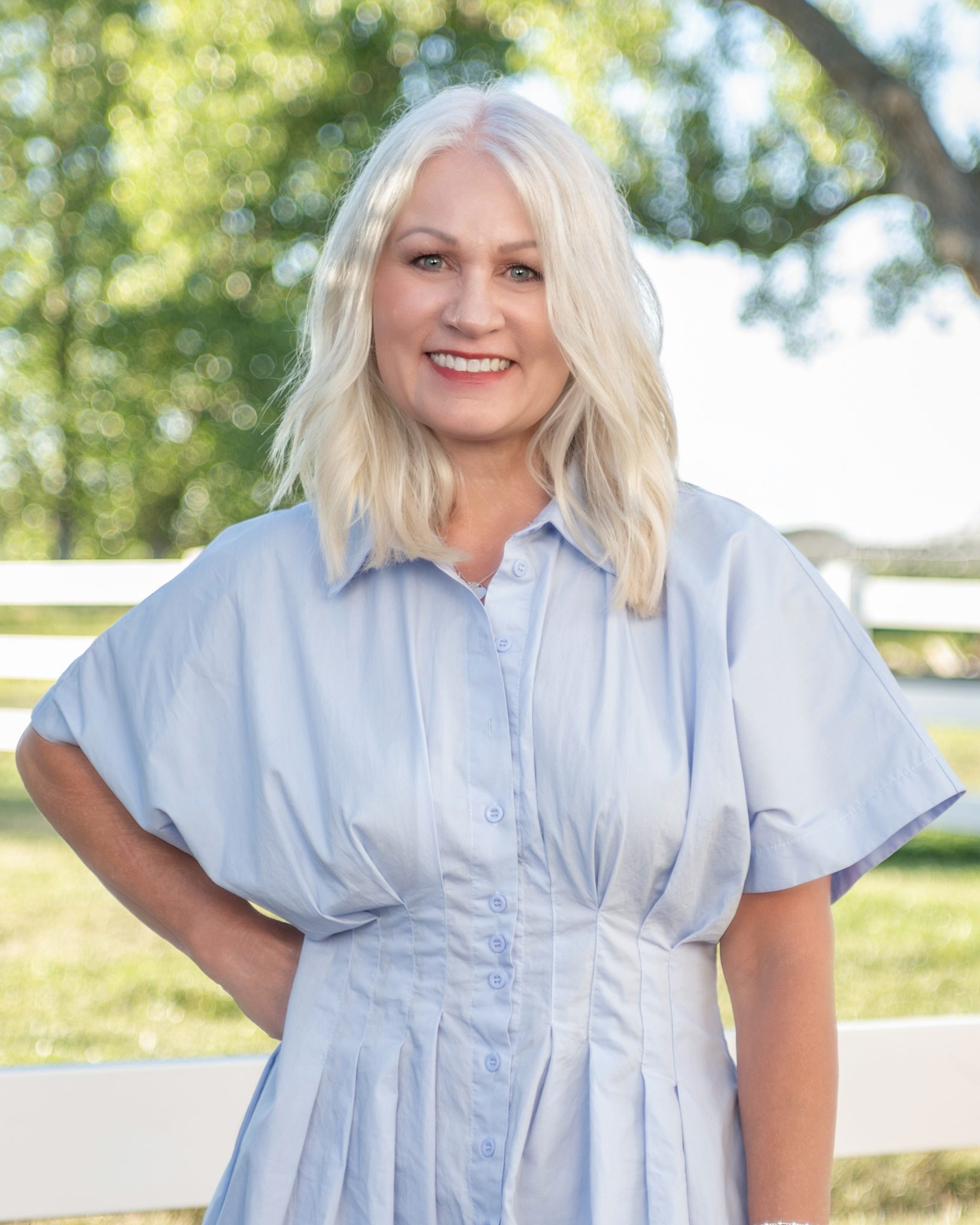 A woman with blonde hair smiling outdoors in a light blue dress.