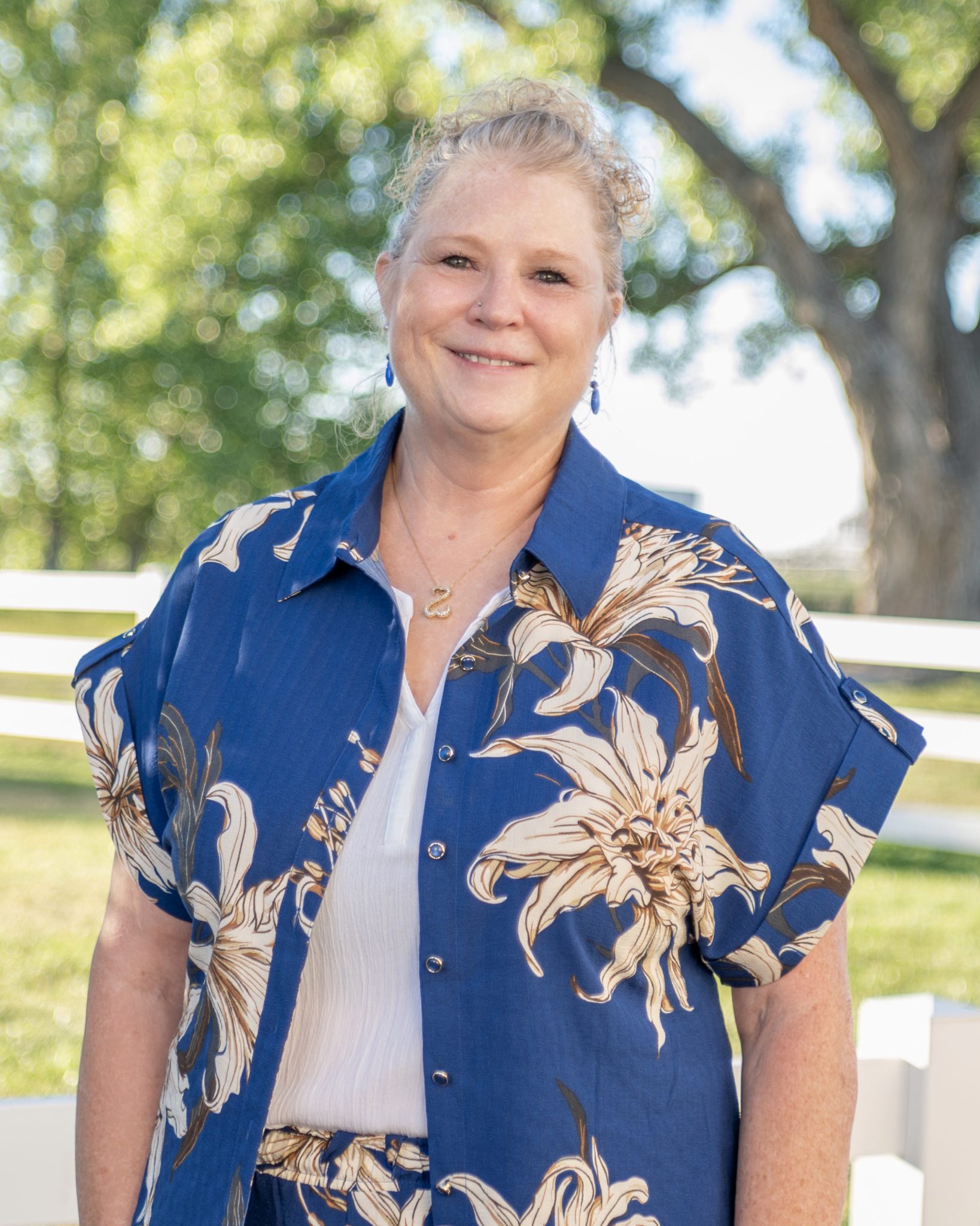 A woman in a blue floral shirt smiles outdoors with trees in the background.