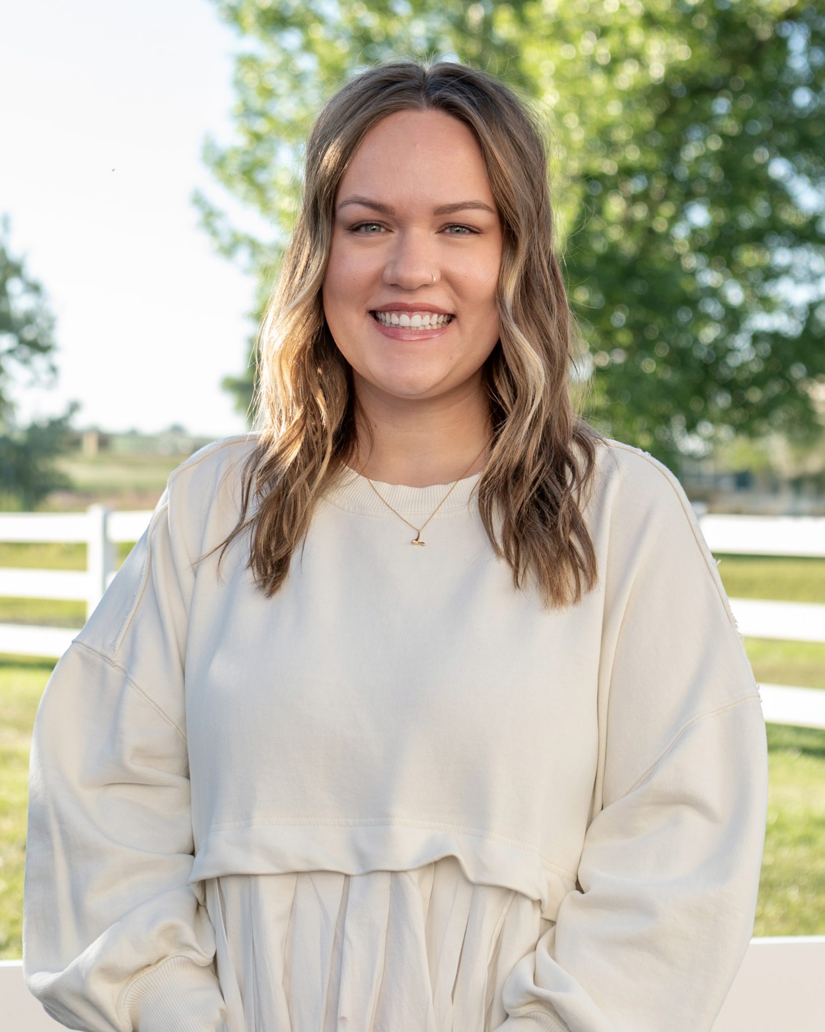 A woman smiling outdoors with trees and a fence in the background.
