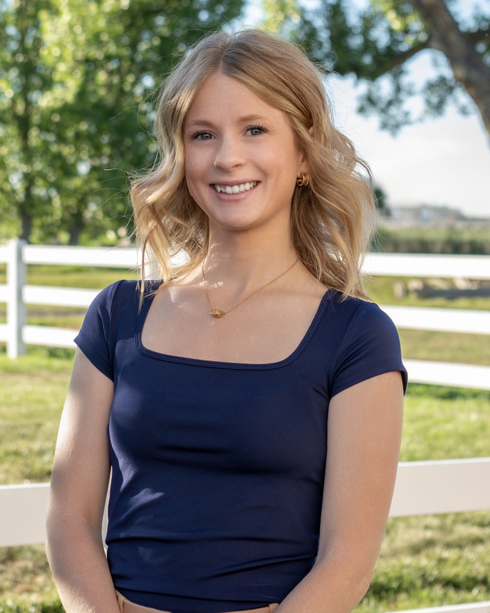 A woman smiling outdoors with trees and a white fence in the background.