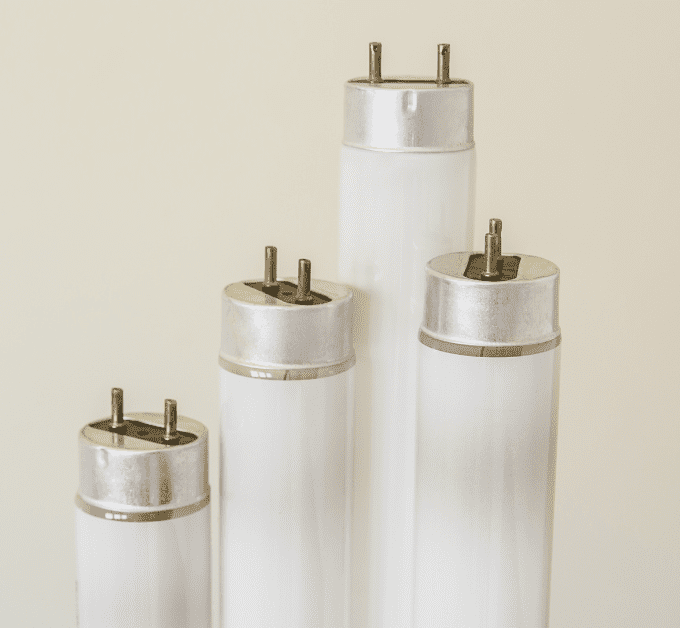 Four fluorescent tube lights of varying lengths stand upright against a plain beige background. Each tube has metal caps with two pins on top, arranged in increasing height from left to right.