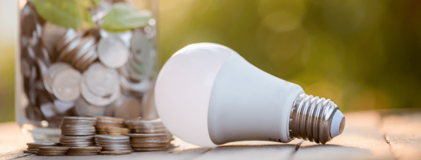 LED bulb on wooden table with stacked coins and jar of coins nearby.