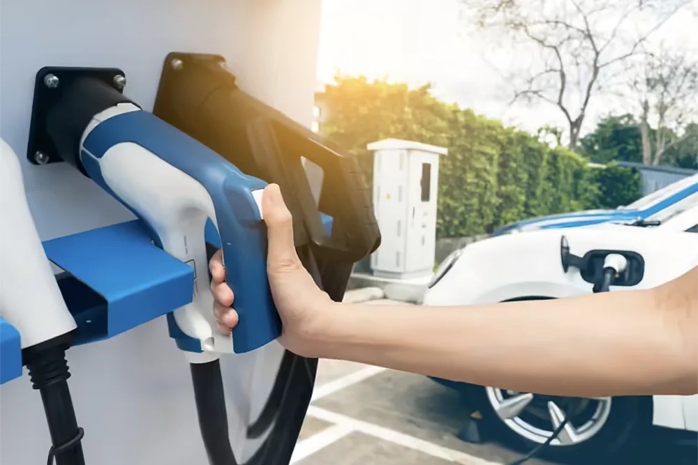 Person charging an electric vehicle at a station with sunlight in the background.