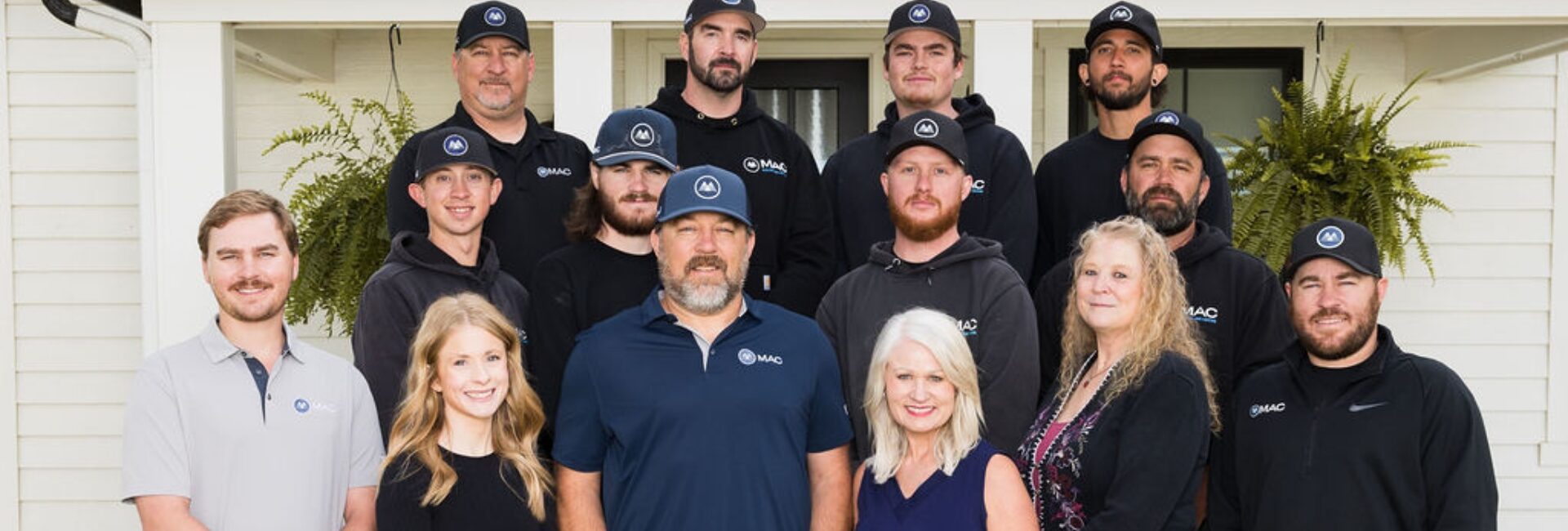 A group of thirteen people, including men and women, stand together outside a white house with green plants by the door. Most of the men wear black shirts and caps; two women and one man are in front, smiling at the camera.