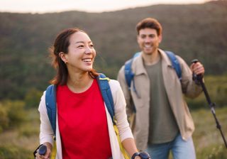 A woman and a man hiking with backpacks and trekking poles in a lush, green landscape.