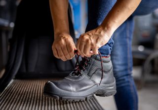 A person tying the laces of black work boots with red accents, on a bench.