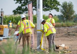 Construction workers install a utility pole at a site, wearing safety vests and helmets.